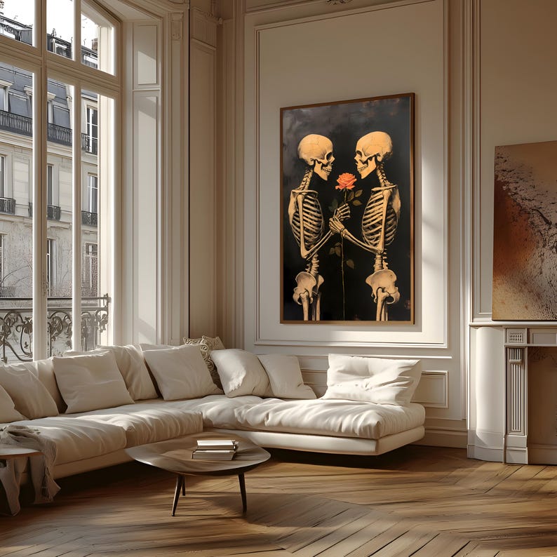 May include: A framed artwork showing two skeletons holding a rose against a dark backdrop. The skeletons are in a loving embrace, with their bony hands gently touching the flower. The artwork is displayed in a well-lit room with a large window and a white sofa.
