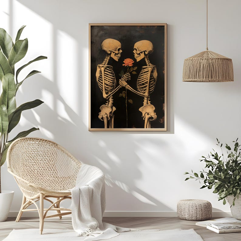 May include: A framed art print depicting two skeletons holding a rose, set against a dark backdrop. The skeletons are rendered in a warm, golden tone. The artwork is displayed in a room with a woven chair and a hanging light fixture.
