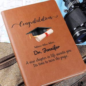 May include: A brown leather journal with the word "Congratulations" in script. A graduation cap and diploma are printed on the cover. The journal is personalized with the name "Dr. Jennifer" and a congratulatory message.