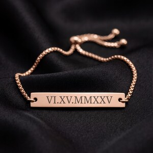 May include: A rose gold-toned bar bracelet featuring a delicate box chain with an adjustable sliding clasp. The central rectangular metal bar is engraved with the Roman numerals VI.XV.MMXXV in a clean, dark, sans-serif font style.