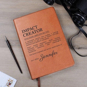May include: A tan leather journal with the words "IMPACT CREATOR" and a definition printed on the cover. The name "Jennifer" is also printed on the cover. A pen and binoculars are in the background.