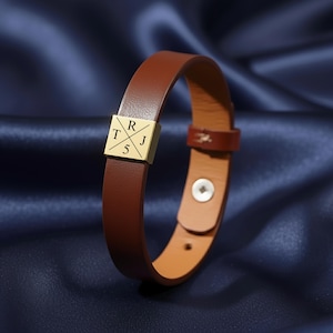 May include: A brown leather bracelet with a gold-colored square accent. The square has the letters "R", "T", "J", and the number "5" engraved on it. The bracelet is fastened with a snap closure.
