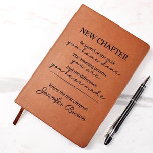 Puede incluir: Un diario de cuero marrón con las palabras "NEW CHAPTER" y texto inspirador. Un bolígrafo negro y plateado descansa junto al diario. El diario incluye el nombre "Jennifer Brown".