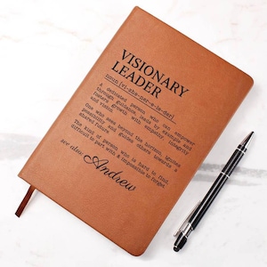 May include: A brown leather journal with the words "VISIONARY LEADER" printed on the cover. The journal includes a definition and the name "Andrew" at the bottom. A black and silver pen rests beside the journal.