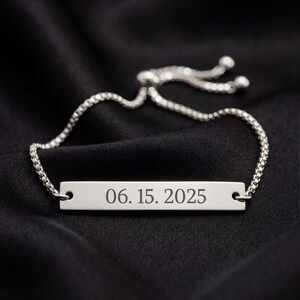 May include: A silver-toned metal bar bracelet featuring a delicate box chain with an adjustable sliding clasp. The rectangular center plate is engraved with the date 06. 15. 2025 in a clear, dark serif font, centered horizontally on the metal surface.