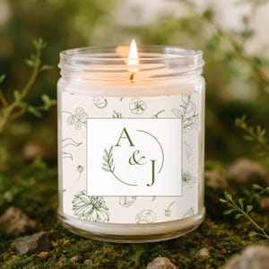 May include: A lit glass jar candle with a white label featuring the initials "A & J" inside a circle, with a green leaf design. The label is surrounded by a floral pattern. The candle sits on a bed of green moss.