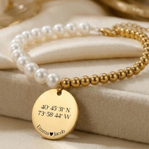 May include: A gold-toned bracelet featuring a combination of white pearls and small, round gold beads. A circular pendant displays engraved coordinates and the names "Emma" and "Jacob" with a heart symbol. The bracelet is displayed on a soft, cream-colored surface.