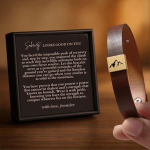 May include: A brown leather bracelet with a gold-colored mountain emblem. The bracelet is held next to a black framed card with the text "Sobriety Looks Good On You" and a message about overcoming challenges.