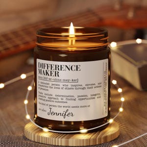 Op de afbeelding: Een brandende bruine glazen kaarsenpot met de tekst "DIFFERENCE MAKER" op een wit etiket. De kaars staat op een houten onderzetter, omringd door lichtslingers. De tekst "Jennifer" is ook zichtbaar.