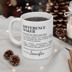 Può includere: Tazza in ceramica bianca piena di liquido scuro, con la scritta "DIFFERENCE MAKER" e una definizione, più il nome "Jennifer". La tazza è circondata da elementi decorativi, tra cui pigne e luci a catena.