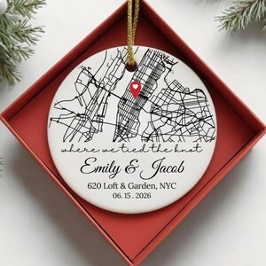 May include: A round white ceramic ornament featuring a black street map print with a red heart-shaped location pin. Text reads: where we tied the knot, Emily & Jacob, 620 Loft & Garden, NYC, 06.15.2026. It hangs from a gold cord inside a red box.
