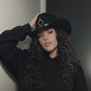 May include: A black cowboy hat with a silver ring detail is worn. The hat is paired with a black hoodie. The model has long, curly brown hair.