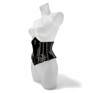 May include: A black corset featuring a glossy finish, vertical rows of metallic gold-toned studs, and a central gold-toned ring detail at the neckline. The garment is displayed on a white mannequin torso, showcasing its structured, pointed hemline.