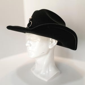 Puede incluir: Un sombrero de vaquero negro con un detalle de anillo de metal plateado. El sombrero tiene un ala ancha y se muestra en una cabeza de maniqu&iacute; blanca. El sombrero est&aacute; hecho de un material suave.