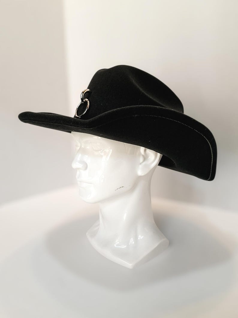 Puede incluir: Un sombrero de vaquero negro con un detalle de anillo de metal plateado. El sombrero tiene un ala ancha y se muestra en una cabeza de maniqu&iacute; blanca. El sombrero est&aacute; hecho de un material suave.