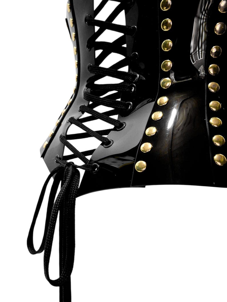 May include: A glossy black corset featuring a vertical row of gold-toned metal studs along the side and a central lace-up closure with black cord. The garment has a structured, reflective surface and a tied cord hanging from the bottom.