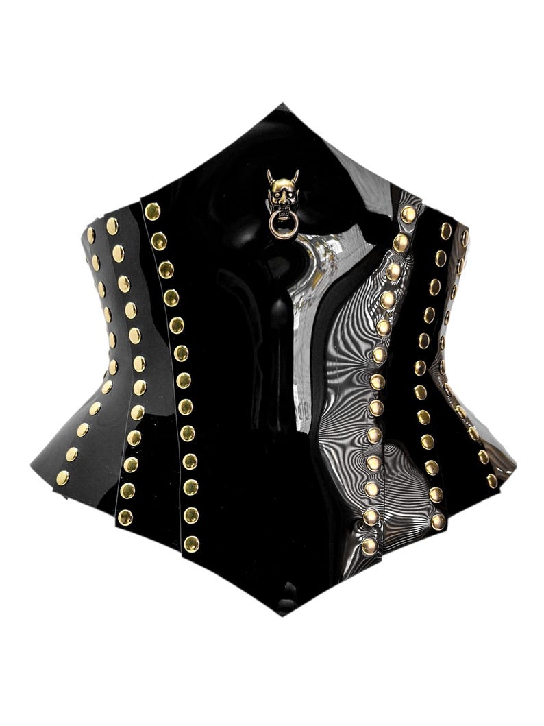 May include: Black corset with gold-colored studs and a devil skull charm. The corset has a glossy finish and a unique shape, with a pointed top and a structured design. The studs are evenly spaced along the edges.