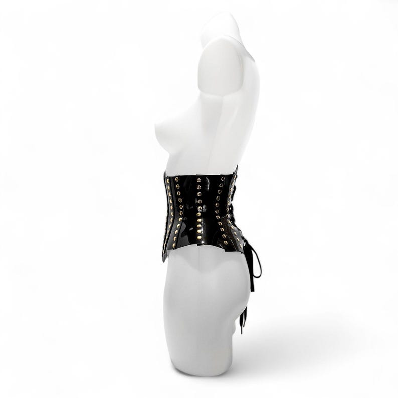 May include: A black glossy corset featuring vertical rows of silver-toned metal eyelets and studs along the front panels. The garment is displayed on a white mannequin torso and includes black lace-up detailing at the back side.