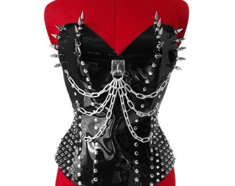 May include: A black glossy strapless corset featuring silver-toned metal spikes along the neckline and sides. The garment is adorned with a central silver metal ring attachment connected to multiple draped silver chains and rows of small metal studs.