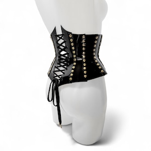 May include: A black corset with gold-colored studs and a lace-up front. The corset is made of a shiny material and is displayed on a white mannequin. The corset has a unique shape and design.