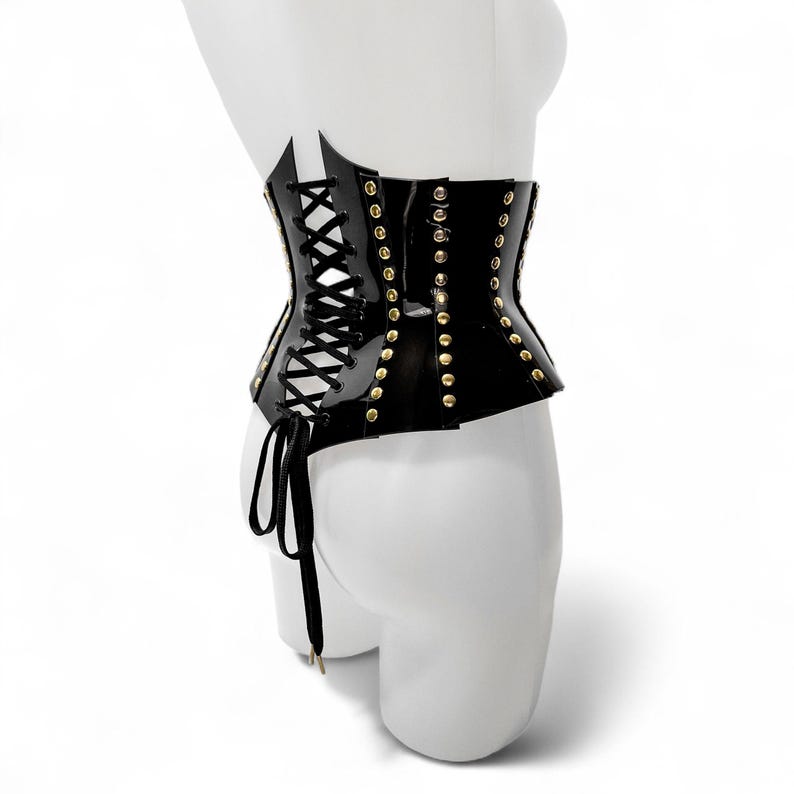 May include: A black corset with gold-colored studs and a lace-up front. The corset is made of a shiny material and is displayed on a white mannequin. The corset has a unique shape and design.