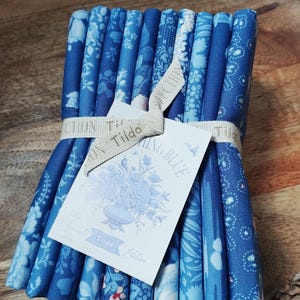 Pode incluir: Uma pilha de tiras de tecido dobradas em vários tons de azul com padrões florais e geométricos. O tecido é amarrado com uma fita bege estampada com a palavra Tilda e apresenta uma etiqueta branca com Tilda Special Edition Spring Blue.
