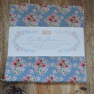 Könnte beinhalten: Ein Stoff-Charm-Pack mit einem Blumenmuster in Rosa und Rot auf hellblauem Hintergrund. Das Paket trägt die Aufschrift "Creating Memories" und enthält 32 Teile, jeweils 12,7 x 12,7 cm. Das Thema ist "Summer and Ocean Blues".