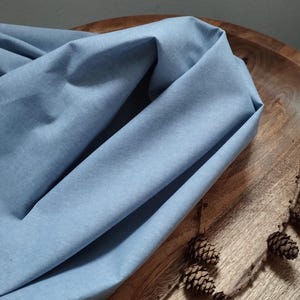 Op de afbeelding: Een lichtblauwe stof gedrapeerd over een houten oppervlak. De stof heeft een subtiele textuur en een gladde afwerking. Een tak met dennenappels bevindt zich op de voorgrond.