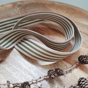May include: A length of striped ribbon in olive green and cream, arranged in a loose, looping pattern. The ribbon is lying on a wooden surface, with a sprig of pinecones in the foreground. The ribbon's design is a classic, versatile pattern.