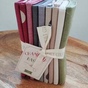 Pode incluir: Uma pilha de quadrados de tecido dobrados em tons de vermelho, cinza, bege e verde, amarrados com uma fita cor de creme. Uma etiqueta de papel retangular está presa à fita, com o texto "CHAMELEON BASICS" e "Tilda" em tinta vermelha e preta.