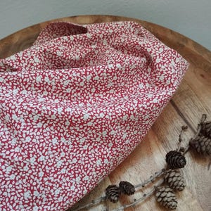 Könnte beinhalten: Ein roter Stoff mit weißem Blumenmuster ist über einem Holztablett drapiert. Der Stoff ist dicht mit kleinen weißen Blüten bedeckt. Mehrere Tannenzapfen und Zweige sind auf dem Tablett angeordnet.