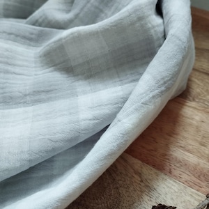 Könnte beinhalten: Ein hellgrauer, strukturierter Stoff ist über eine Holzoberfläche drapiert. Der Stoff hat ein dezentes Karomuster und wirkt weich. Zwei Tannenzapfen und ein Zweig befinden sich im Vordergrund und fügen der Komposition ein natürliches Element hinzu.