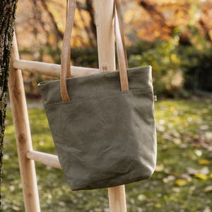 May include: Olive green tote bag with tan leather handles, hanging on a wooden ladder. The bag has a simple design with a front pocket. The background is a blurred view of a park with trees and foliage.