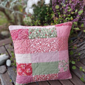 Könnte beinhalten: Quadratisches Patchwork-Kissen mit verschiedenen roten, rosa und grünen Blumen- und Streifenmustern. Das Kissen ist gesteppt und hat ein weiches, strukturiertes Aussehen. Es liegt auf einer Holzoberfläche.