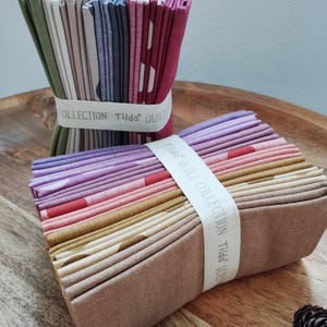 Può includere: Due pile di quadrati di tessuto piegati, ciascuna fissata con una fascia di carta bianca etichettata Tilda Quilt Collection. La prima pila presenta toni verdi, grigi, blu e magenta. La seconda mostra sfumature viola, rosa, oro e marrone chiaro.