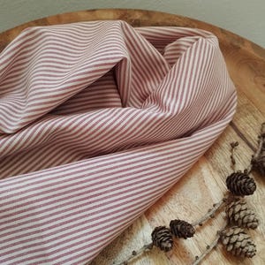 Pode incluir: Close-up de um tecido dobrado com um padrão de listras vermelhas e brancas. O tecido está disposto sobre uma superfície de madeira, com um ramo de pinhas em primeiro plano. A textura do tecido é visível.