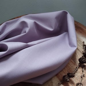 Könnte beinhalten: Ein weicher, helllilafarbener Stoff, der über eine Holzoberfläche gelegt ist. Der Stoff hat eine glatte Textur und einen dezenten Glanz.