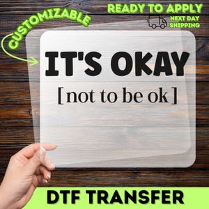 May include: A clear DTF transfer sheet featuring the black text IT'S OKAY [not to be ok] in a bold, sans-serif font for the first line and a smaller, serif font for the second line, presented on a transparent rectangular film.