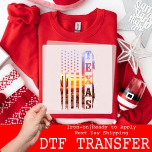 May include: A red crew neck sweatshirt with a DTF transfer featuring a Texas flag design. The flag design includes stars, a sunset, and the word "TEXAS". The image also includes a Christmas stocking, a Santa-themed wine glass, and a star-shaped decoration.