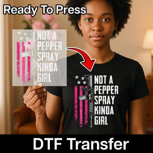 May include: Black t-shirt with the text "NOT A PEPPER SPRAY KINDA GIRL" and a graphic of a rifle and American flag design. The image also shows a DTF transfer and the words "Ready To Press".