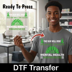 May include: A gray t-shirt featuring a green sunflower graphic with the text THE SUN WILL RISE AND WE WILL TRY AGAIN MENTAL HEALTH AWARENESS and two green ribbons. A matching DTF transfer sheet is held up, showing the same design.