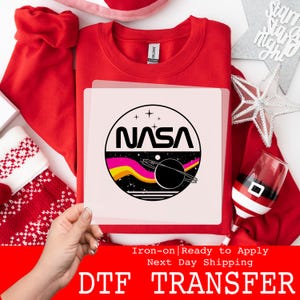 May include: A red crew neck sweatshirt with a NASA logo iron-on transfer. The NASA logo features a black planet with a ring, a yellow and pink wave, and the word "NASA" in white. The transfer is ready to apply.