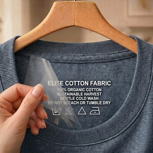 Peut inclure: Un t-shirt gris foncé sur un cintre en bois. Le t-shirt porte une étiquette avec le texte "ELITE COTTON FABRIC", "100% ORGANIC COTTON" et des instructions d'entretien. Une main tient l'étiquette.