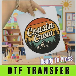 May include: A white DTF transfer sheet featuring a circular graphic with a retro sunset design in orange, yellow, and teal. The text "Cousin Crew" is printed in a black script font over a silhouette of pine trees and mountain peaks.