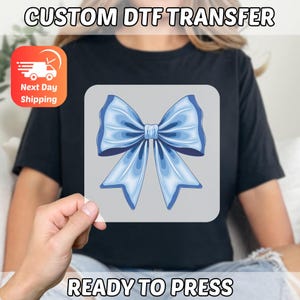 May include: A light blue bow design on a gray background, ready to press. The text "CUSTOM DTF TRANSFER" and "READY TO PRESS" are displayed. An orange and white graphic with the words "Next Day Shipping" is also visible.
