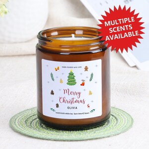 Può includere: Una candela in barattolo di vetro marrone con un'etichetta bianca che recita "Merry Christmas" e "OLIVIA". L'etichetta presenta illustrazioni a tema natalizio. La candela è su un sottobicchiere verde. Una stella rossa dice "MULTIPLE SCENTS AVAILABLE".