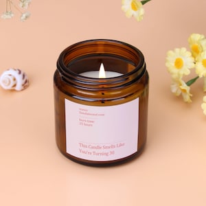 Peut inclure: Un pot de bougie en verre brun allumé avec une étiquette blanche qui dit "This Candle Smells Like You're Turning 30." L'étiquette indique également que le parfum est de rose de santal et le temps de combustion est de 25 heures. La bougie est entourée de fleurs.