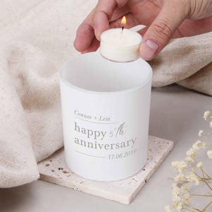 May include: A white candle holder with the text "Connor + Leia happy 5th anniversary 17.06.2019" is shown. A lit tea light candle is held above the holder. The candle holder sits on a square, textured coaster.