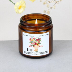 Può includere: Una candela accesa in un barattolo di vetro marrone con un'etichetta floreale "Get Well Soon". L'etichetta presenta un bouquet di fiori di vari colori. La candela ha la fragranza "Blue Wind Chime" e un tempo di combustione di 16 ore.