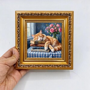 May include: A framed oil painting depicting an orange tabby cat sleeping on a stack of three books. The scene includes a blue and white checkered tablecloth and a glass vase filled with pink roses, all encased in an ornate gold frame.